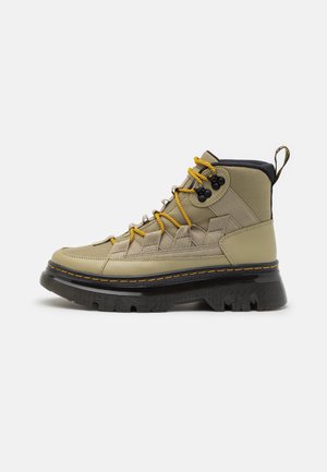 Botte haute kaki avec semelle noire épaisse, lacets jaunes, œillets noirs et languette au talon, vue de profil sur fond blanc.