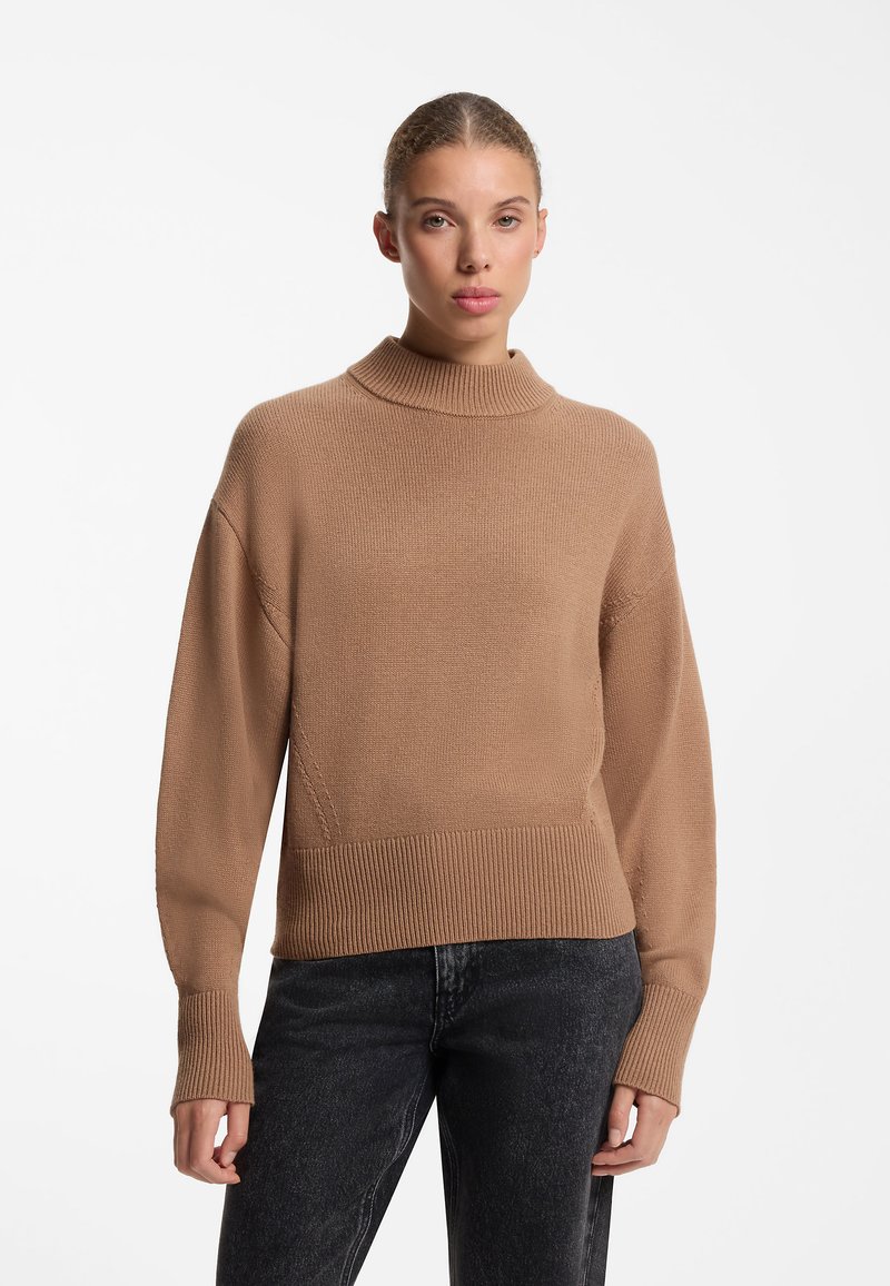Jeune femme aux cheveux attachés, portant un pull en maille marron et un jean foncé, debout devant un fond blanc.