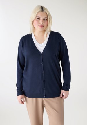 Giovane donna con capelli chiari che indossa un cardigan blu navy con bottoni, top bianco e pantaloni beige su uno sfondo bianco semplice.