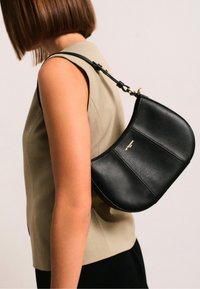 Borsa a spalla in pelle nera con design curvo, dettagli in oro e finitura texture, indossata su una cima beige contro uno sfondo neutro.