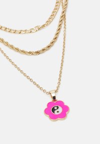 Colar de corrente em camadas douradas com um pendente em forma de flor rosa, apresentando um símbolo Yin Yang em preto e branco no centro.