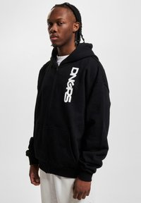 Hoodie noir zippé en tissu doux avec une coupe relaxée. Il présente un logo blanc sur le côté gauche et des poches avant.