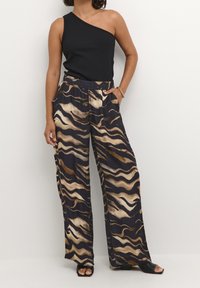 Nainen, joka on pukeutunut mustaan yhden olkapään yläosaan ja avariin housuihin, joissa on abstrakti beigen ja ruskean aaltoileva kuvio. Asusteena mustat avointoepäritiiliset korkokengät.