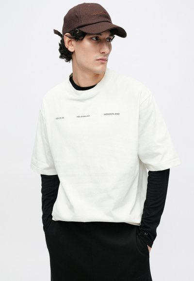 Biela oversize tričko vrchná vrstva cez čierne tričko s dlhým rukávom. Vyšívaná hnedá čiapka s logom "HOLZWEILER". Jednoduchý, pohodlný dizajn.