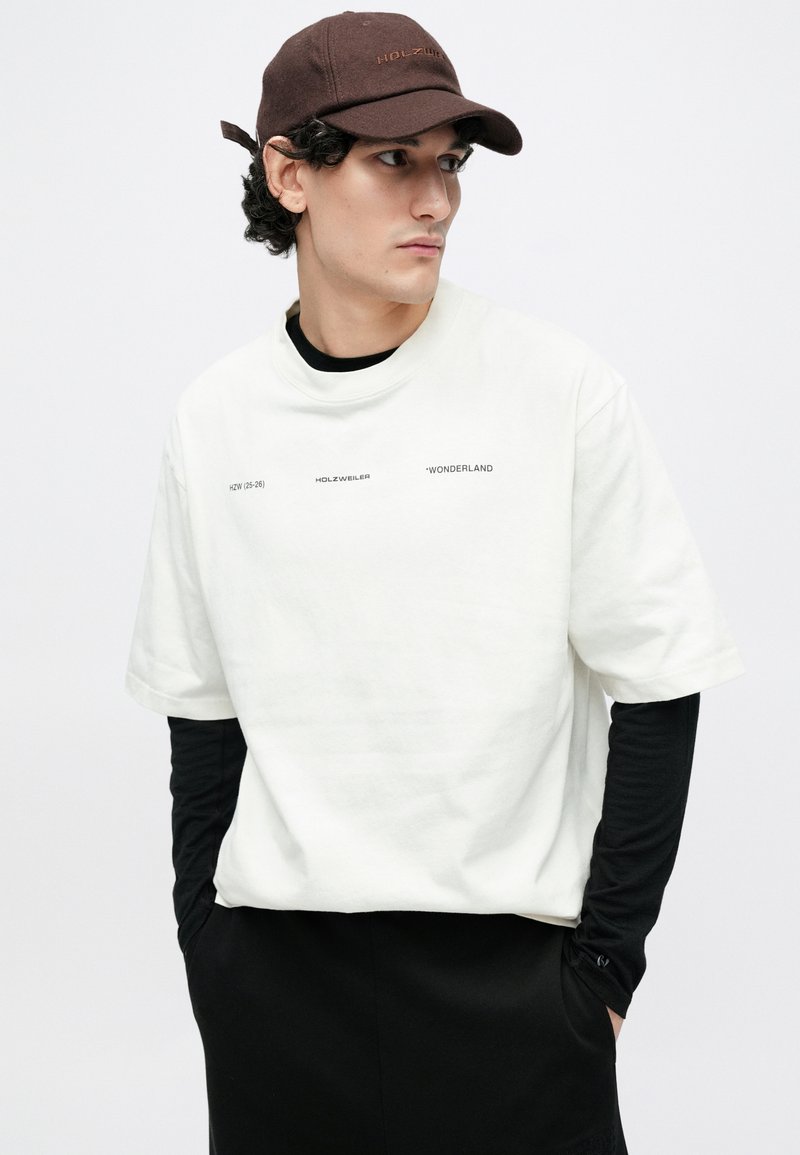 Maglietta bianca oversize indossata sopra una camicia nera a maniche lunghe. Cappellino marrone ricamato con il logo "HOLZWEILER". Design semplice e rilassato.