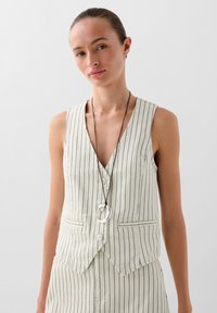  Lichtbeige gestreepte vest met diepe V-hals, voorzien van knopen en twee zakken. Gecombineerd met een lange ketting met ronde accenten.