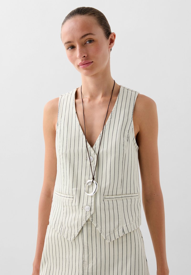  Lichtbeige gestreepte vest met diepe V-hals, voorzien van knopen en twee zakken. Gecombineerd met een lange ketting met ronde accenten.