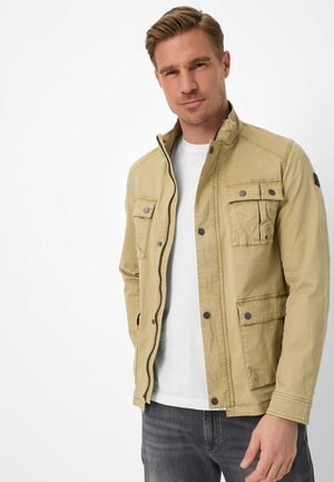 Man draagt een beige jasje met knopen over een wit overhemd en donkere spijkerbroek, staand tegen een effen witte achtergrond.