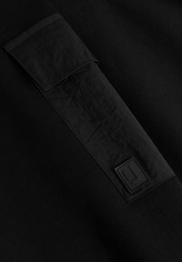 Felpa nera con un distintivo a livello della spalla in tessuto testurizzato e un'etichetta con il logo. Tessuto liscio con dettagli di cucitura sottili in tutto il capo.