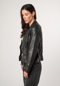 Schwarze Lederbikerjacke mit tailliertem Schnitt, Reißverschluss vorne und markanten Nähten. Über einem weißen Oberteil getragen und kombiniert mit dunklen Jeans.