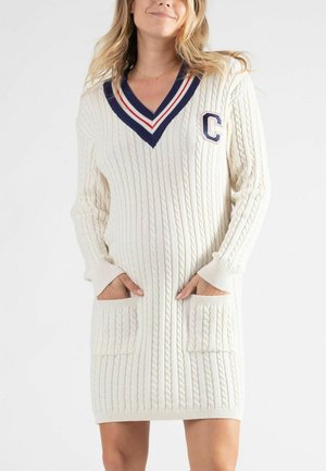 Robe pull en crème avec motif en cable. Présente un décolleté en V avec des bordures bleu marine et rouge, un patch logo, et deux poches avant. Manches longues.