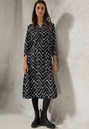 Schwarz-weißer, mit Zickzackmuster versehener Kleid aus glattem Stoff, mit Kragen und Dreiviertelärmeln, kombiniert mit schwarzen Ankle Boots.
