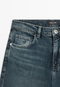 Tmavě modré džíny z denimu s přední kapsou, kovovým zapínáním na knoflík a štítkem s nápisem "Massimo Dutti". Měkká textura a klasický design.
