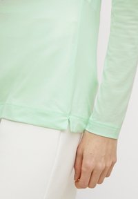 Camicia a maniche lunghe verde chiaro in tessuto morbido, con orlo arrotondato e spacchi laterali, indossata sopra pantaloni bianchi.
