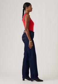 Γυναίκα με πλεξούδες που φορά κόκκινο αμάνικο τοπ, σκούρα μπλε τζιν ψηλόμεσα, μαύρα μπότες με τακούνι και ασημένια κοσμήματα, όρθια σε πλάγια όψη.