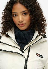 Hvid vandtæt jakke med sorte lynlåse, der har en mørk navy ribbet højhalset trøje. Viser et logo patch på brystet. Krøllet hår.