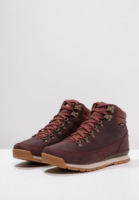 Brązowe i bordowe buty trekkingowe z skórzanym wierzchem, metalowymi elementami, teksturowaną gumową podeszwą i wyściełanym kołnierzem na kostkę. Wąskie sznurowadła z oczkami.