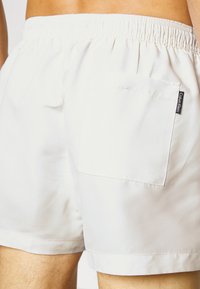 Pantaloni da bagno beige chiaro con vita elastica e tasca sul retro. Texture liscia con un design minimalista e un'etichetta di branding discreta.