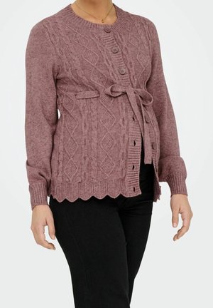 Frau trägt einen mauvefarbenen, kabelgestrickten Cardigan mit Knöpfen und gebundenem Gürtel über schwarzen Hosen, gezeigt vom Hals bis zu den Hüften.