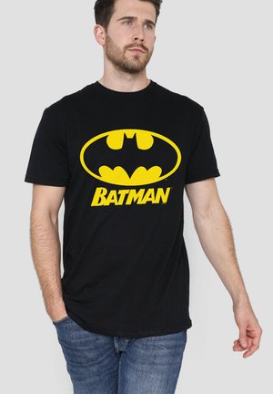 Man wearing black t-shirt with large yellow Batman logo and text, paired with blue jeans, standing against a plain background.