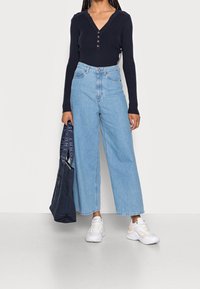 Haut côtelé bleu marine à manches longues avec un col en V et des boutons, associé à un jean large bleu clair et des baskets blanches. Sac fourre-tout à la main.