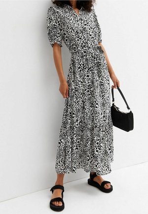 Kobieta w długiej sukni w czarno-białe cętki z bufkami na rękawach, trzymająca małą czarną torebkę i nosząca czarne sandały.