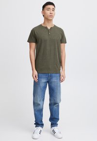 Oliven grønn kortermet henley-skjorte med treknappslukking, sammen med lys blå rettbukser og hvite sneakers med svarte striper.