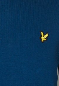 Maglione blu lavorato a maglia con una texture a coste, caratterizzato da un piccolo emblema d'aquila dorato sul lato sinistro. Il tessuto sembra morbido ed elastico.