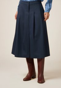 Gonna plissettata blu navy realizzata in tessuto misto lana con chiusura a bottone, abbinata a stivali alti al ginocchio in pelle marrone. Texture liscia, forma a campana.