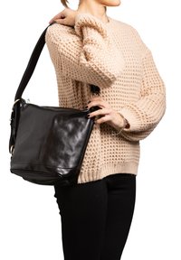 Borsa a spalla in pelle nera con una forma rettangolare, texture morbida e tracolla regolabile, tenuta accanto a un maglione beige lavorato a maglia.