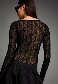 Čierne čipkované body s dlhými rukávmi, s kvetinovým vzorom a priehľadným chrbtom. Kombinované s textúrovanou čiernou sukňou.