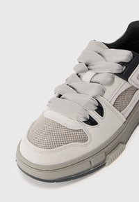 Scarpa sportiva grigio chiaro realizzata in mesh e suede, con superficie texturizzata, lacci piatti e una suola in gomma durevole con accenti a contrasto.