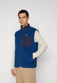 Blauwe fleece vest met hoge kraag en volledige rits. Heeft een donkerblauwe borstzak en zijzakken met rits. Draag het over een licht shirt met lange mouwen.