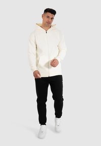 Weißer Kapuzenpullover mit Reißverschluss und beigem Innenfutter, kombiniert mit schwarzen, taillierten Jogginghosen und weißen Turnschuhen. Der Kapuzenpullover verfügt über eine Kapuze mit Kordelzug und Taschen.