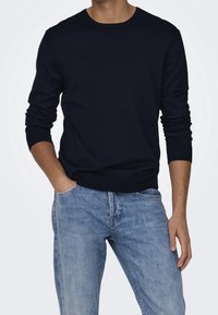 Pull en tricot bleu marine à manches longues et col rond, associé à un jean en denim bleu clair. Le matériau semble lisse et ajusté.