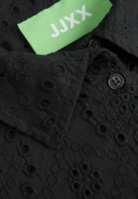 Cuello de camisa bordado en negro con patrones circulares; presenta una etiqueta verde con "JJXX" y dos botones negros. Textura suave.