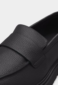 Μαύρο μοκασίνι από δερμα με υφή, με ραμμένη ταινία και χοντρή σόλα, που δείχνει κοντινή εικόνα του άνω μέρους και του ανοίγματος του παπουτσιού.