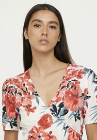 Robe fleurie avec un décolleté en V. Présente des motifs floraux rouges, roses et bleus sur un fond crème. Manches courtes bouffantes, texture de tissu douce.