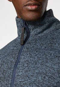 Dark blue zip-up fleece jacket with a textured pattern. Features a high collar and a black zipper pull for closure.
