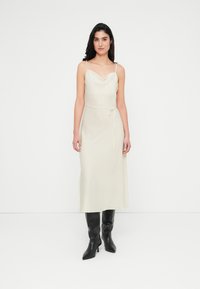 Midi φόρεμα χωρίς μανίκια με λαιμόκοψη καμπούρας σε ανοιχτό κρεμ ύφασμα. Διαθέτει ζώνη δεσίματος στη μέση, μαλακή υφή και αέρινη σιλουέτα. Φορεμένο με μαύρες μπότες μέχρι το γόνατο.