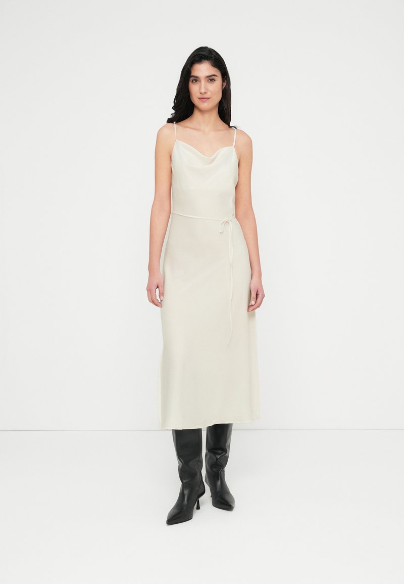 Midi φόρεμα χωρίς μανίκια με λαιμόκοψη καμπούρας σε ανοιχτό κρεμ ύφασμα. Διαθέτει ζώνη δεσίματος στη μέση, μαλακή υφή και αέρινη σιλουέτα. Φορεμένο με μαύρες μπότες μέχρι το γόνατο.