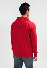 Felpa rossa con cappuccio e maniche lunghe, caratterizzata da un design semplice e una texture morbida, abbinata a jeans in denim blu.