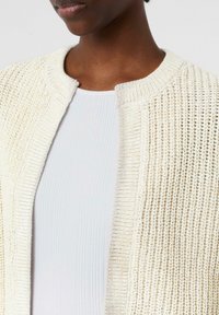 Cardigan en tricot de couleur écrue avec une finition texturée, présentant un col rond et un design à devant ouvert, porté sur un haut côtelé clair.