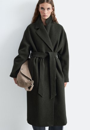 Femme portant un long manteau ceinturé vert foncé tenant un sac à main en daim beige, debout devant un mur clair uni.