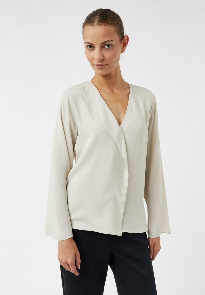 Blusa crema con scollo a V e maniche lunghe, realizzata in tessuto morbido. Presenta un design frontale drappeggiato e una leggera lucentezza. Abbinata a pantaloni scuri.