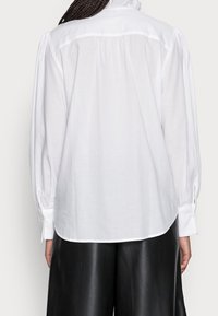 Camicia bianca a maniche lunghe con schiena arricciata, tessuto liscio, polsini con bottoni e vestibilità rilassata. Abbinata a pantaloni neri lucidi.