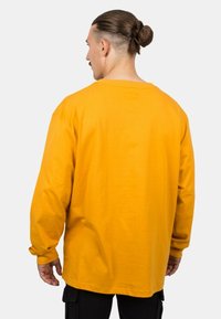 Langarmshirt aus leuchtend gelbem Baumwollstoff; lockere Passform mit abgesenkten Schultern und einem Rundhalsausschnitt. Nahtdetails und gerippte Bündchen sind sichtbar.