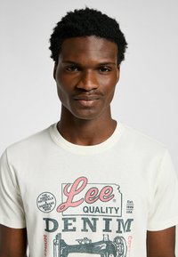 Camiseta de algodón color crema con un estampado gráfico de estilo vintage en rosa y negro, con el texto "Lee Quality Denim" y una ilustración de máquina de coser.