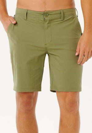 Mężczyzna stojący, ubrany w oliwkowozielone krótkie spodenki do kolan z zapięciem na guziki i suwak, ręce swobodnie opuszczone wzdłuż ciała.