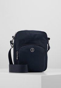 Kompaktowa granatowa torebka crossbody z przednią kieszenią na zamek, srebrnym logo "B", regulowanym paskiem i prostokątnym kształtem na białej powierzchni.